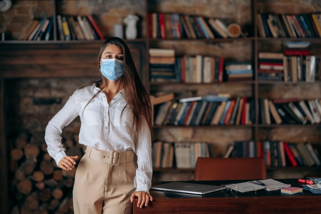 Young woman in medical face mask at home on bookshelves background